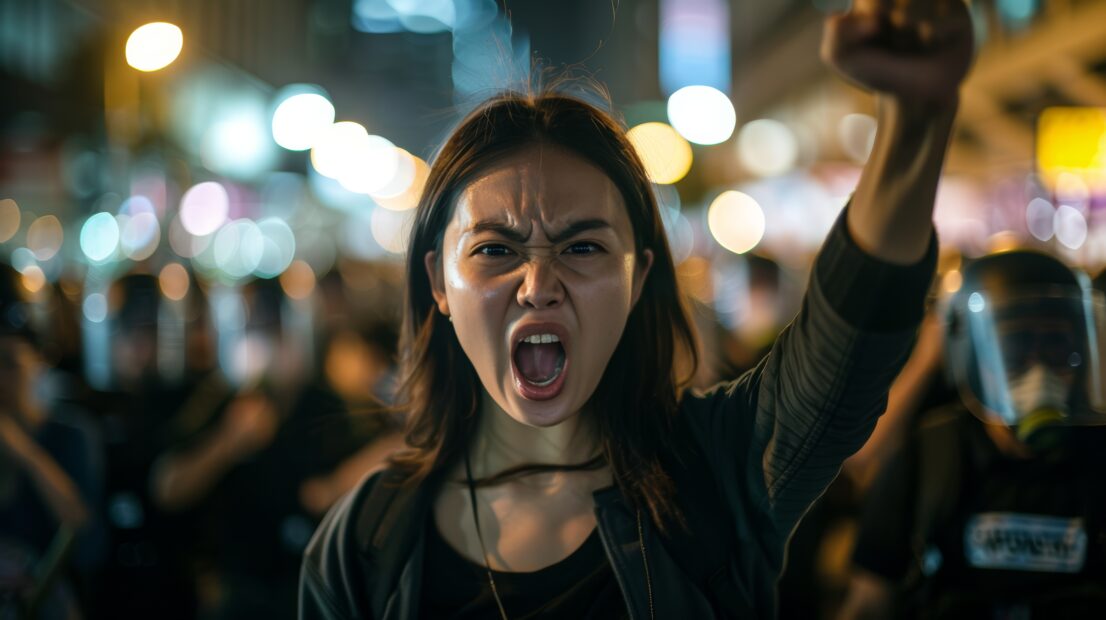 Photograph of a young Asian woman expressing anger during a protest in Hong Kong, her fist raised, against a backdrop of police and demonstrators under overcast, diffuse lighting to convey the emotion and chaos of civil unrest with a stark, realistic color palette, medium shot, influenced by photojournalism. Shot with Nikon D850 and AF-S NIKKOR 35mm f/1.4G lens --chaos 15 --ar 16:9 --style raw Job ID: eb7b1a9d-7e7b-4f5b-9d78-3933b45bc569